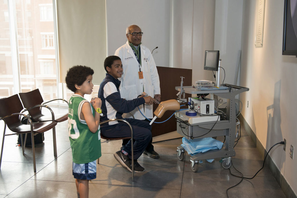 Teen boy demontrates minimally invasive knee surgery while older man and young boy look on in MGH Museum