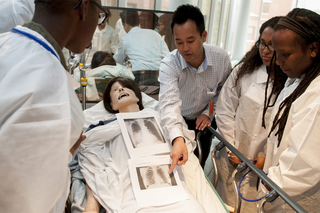 Simulation mannequin lies on a table while man points to xray scans and three people look on