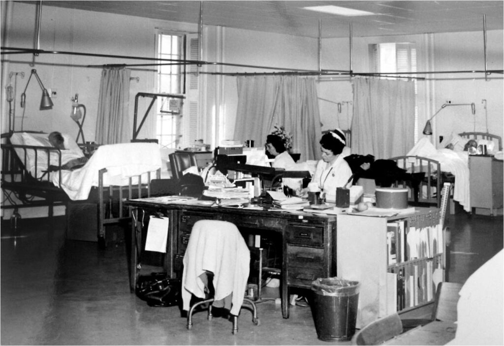 black & white photo of two nurses at a desk in the center and two patients in bed on thee left and right behind them.
