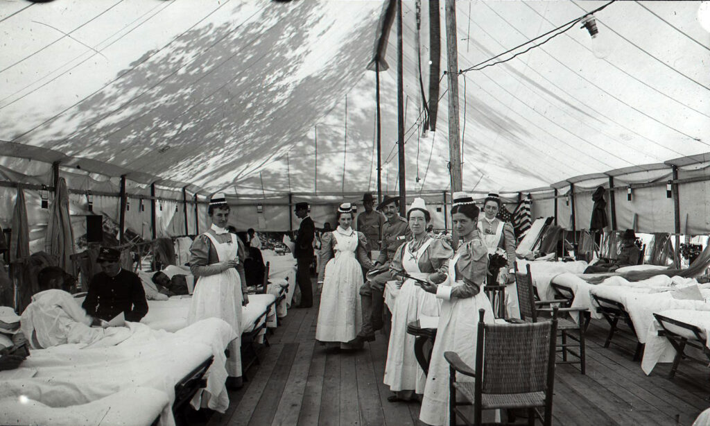black & white photo of several nurses and bed-ridden patient in a large tented room