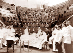 sepia-toned photo of several men in white coats attending an operation, with many more men in the theater rows above observing