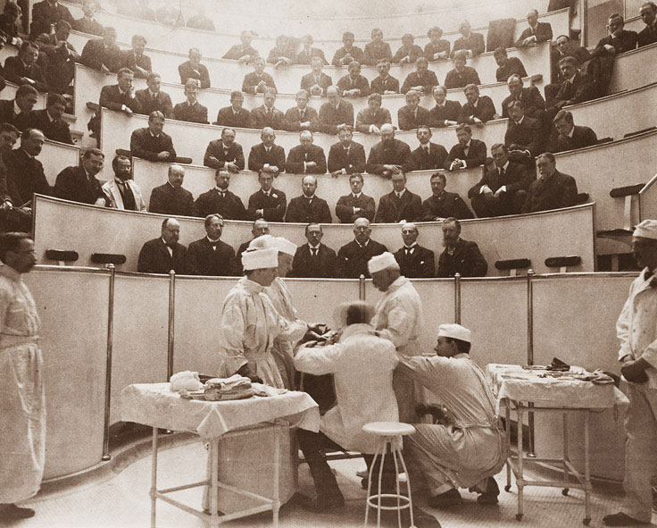 sepia-toned photo of eight men in white coats attending an operation, with several dark-suited men in the theater rows above observing