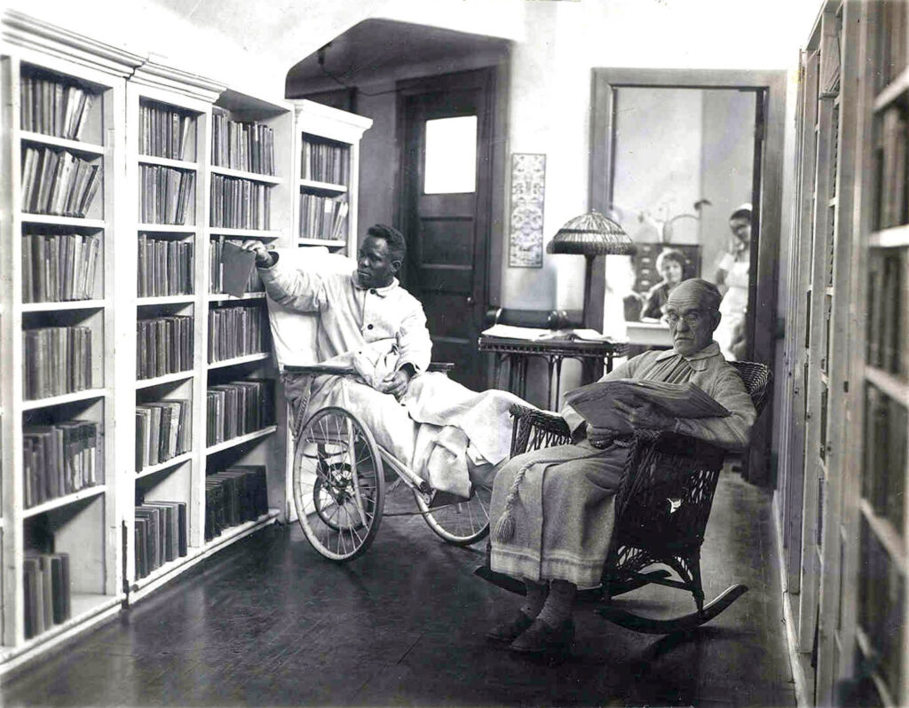 black & white photo of two men in a library. The man on the left is in a wheelchair, reaching for a book, and the man in the right reading in a rocking chair