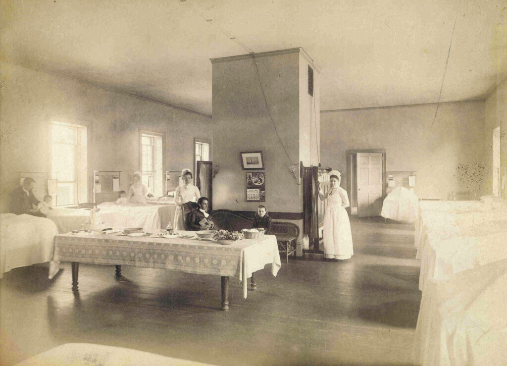 A room with a table, chairs, and beds, with adults and children.