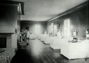 black & white photo of large, sunlit room with eighty beds in view with four nurses and five patients