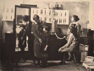 black & white photo shows four people, including a woman examing a girl's posture in a mirror.