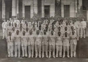 black & group photo of several rows of people standing in white uniforms. Each person's last name is written on their chests