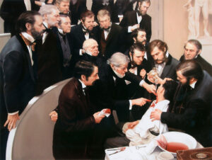 Several dark suited men observing a surgery on a seated, unconscious patient performed by a man with white hair and beard.