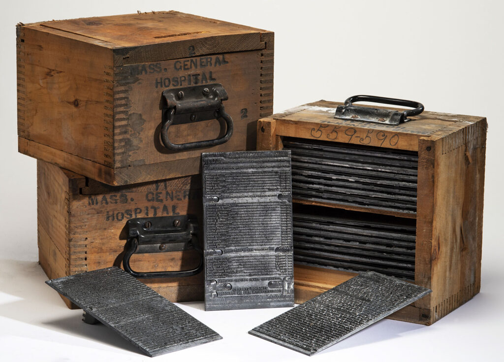 Three small wooden boxes with metal handles. One is opened to display a stack of metal printing plates, three of which are propped up in front of the boxes